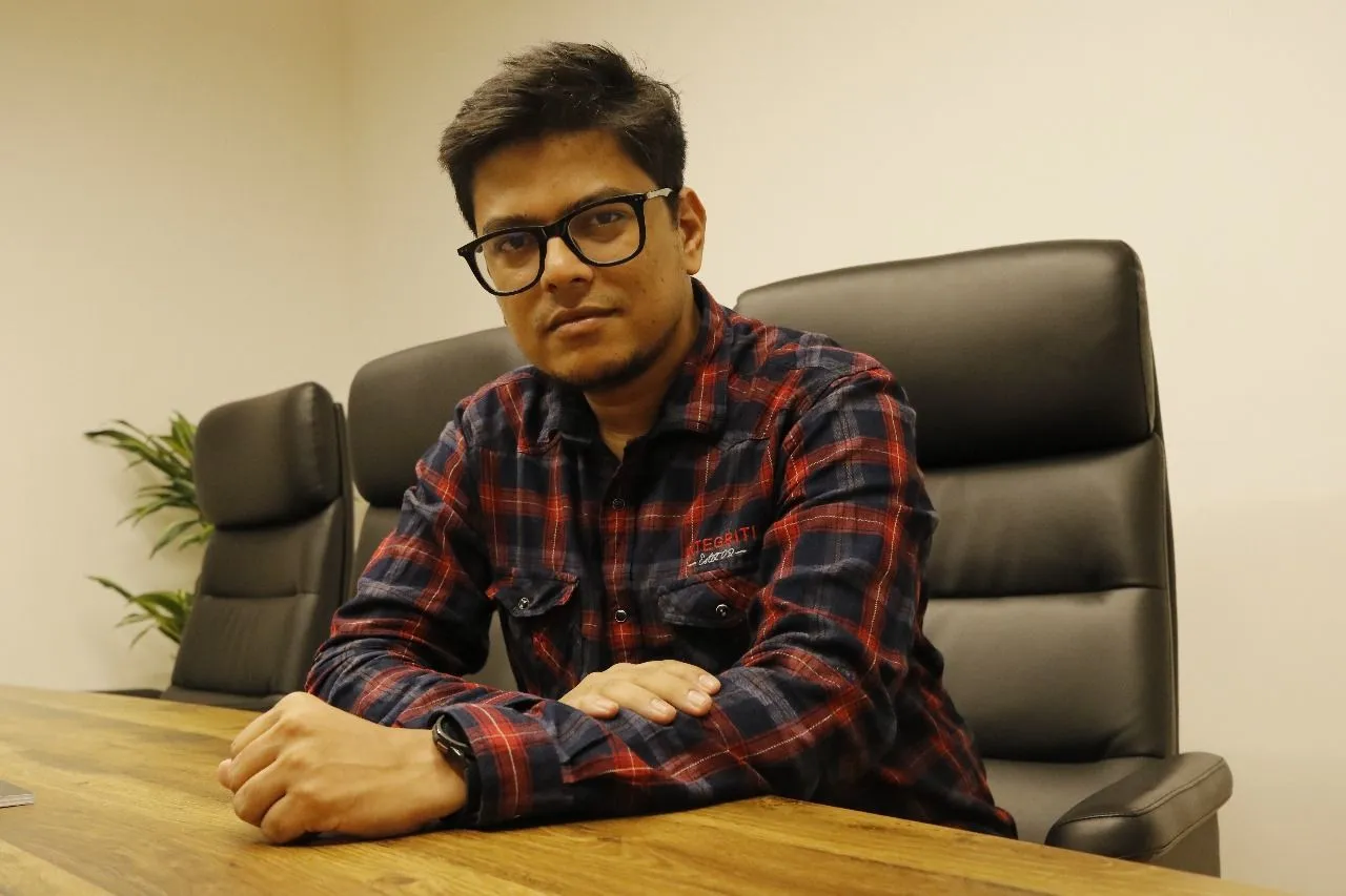 A man wearing glasses sits at a table in a modern office, focused and engaged in his work.