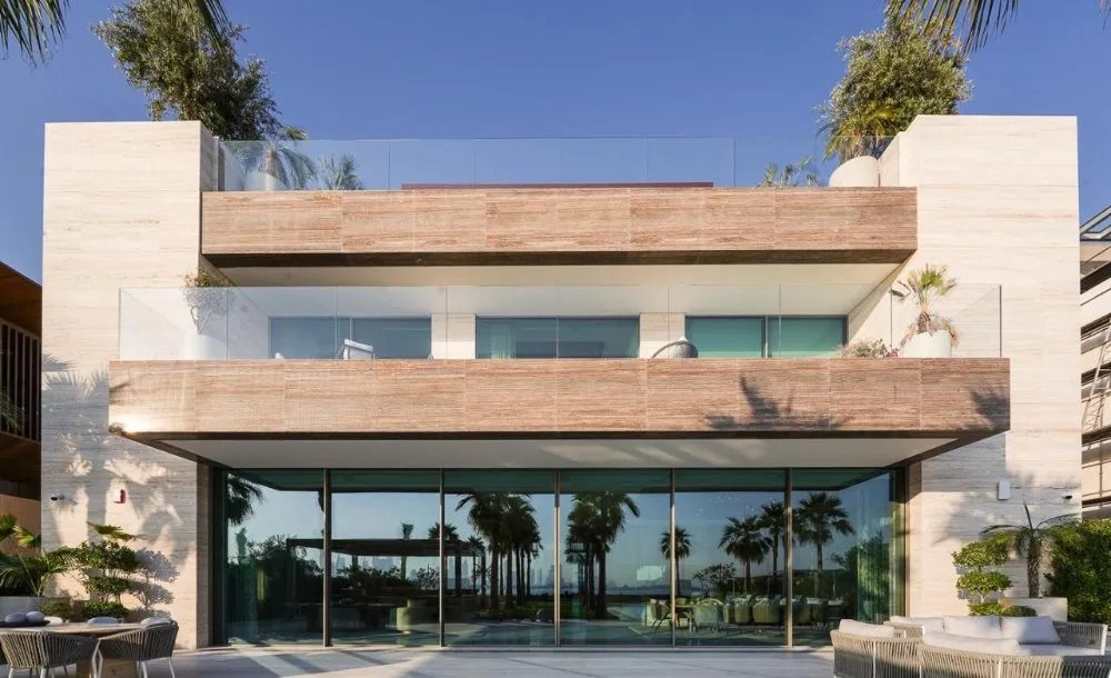 Modern coastal villa with wooden accents, glass balconies, and palm trees, reflecting luxury and elegance against a clear blue sky.