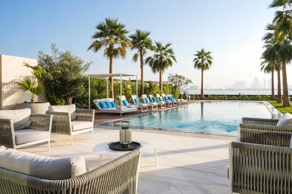 Luxurious outdoor pool area with palm trees, sun loungers, and modern seating, offering a tranquil retreat with a city skyline in the background.