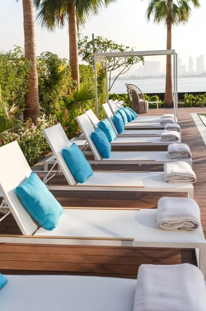 A row of white lounge chairs with turquoise cushions, surrounded by lush greenery and palm trees, overlooking a serene waterfront view.