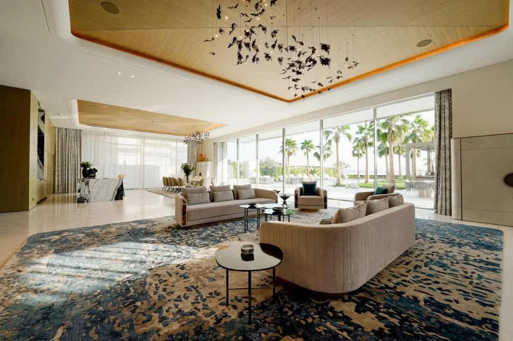 A modern living room with plush seating, a large rug, and a decorative ceiling fixture, featuring palm trees visible through floor-to-ceiling windows.
